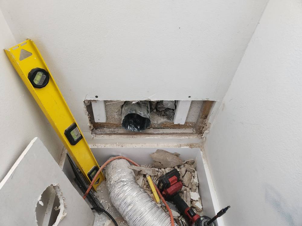 Before: laundry closet wall cut open to expose a disconnected metal dryer vent with drywall debris below